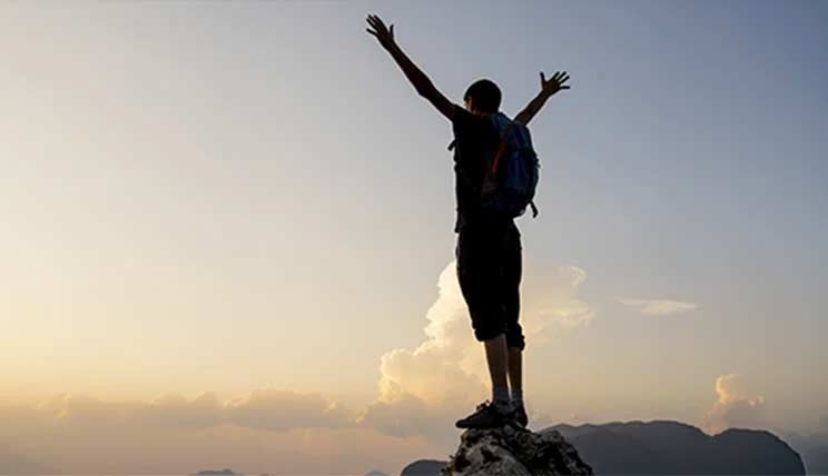 A hiker with a backpack standing on a mountain peak at sunset, arms raised in celebration.